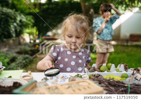 Little girl looking at models of insect with magnifying glass, learning about wildlife in forest kindergarten. 117519350