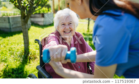 Female caregiver doing motorized exercises with senior woman in wheelchair. Elderly woman working out with small dumbbell, nurse leading and teaching her. Female caregiver doing motorized exercises with senior woman in wheelchair. Elderly woman working out with small dumbbell, nurse leading and teaching her. 117519367