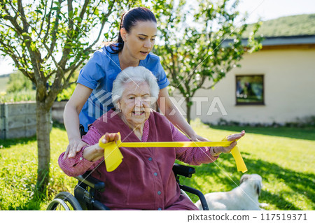 Female caregiver doing motorized exercises with senior woman in wheelchair. Nurse and elderly woman working out with resistance band. Female caregiver doing motorized exercises with senior woman in wheelchair. Nurse and elderly woman working out with resistance band. 117519371