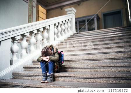 Portrait of bullied girl is crying on the steps in front of the school, waiting for her parents. Student girl with bag after day in elementary school. 117519372