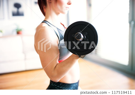 Beautiful redhead woman's home workout routine with dumbbell. Home fitness workout in living room, sporty woman wearing sportswear. 117519411