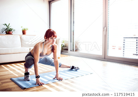 Beautiful redhead woman's home fitness routine. Home workout on gym mat in living room, sporty woman wearing sportswear. 117519415