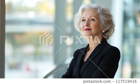 confident older woman dressed in professional attire, standing in a modern office space with large windows 117519416