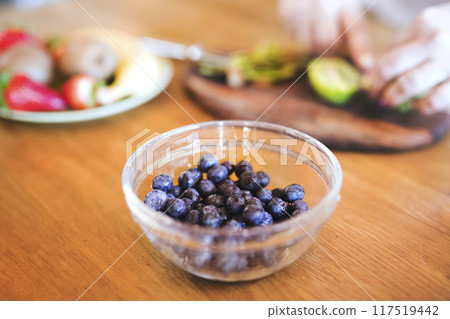 Bowl with fresh blueberries for smoothie. Overweight woman in the kitchen cutting fruit on cutting board. Bowl with fresh blueberries for smoothie. Overweight woman in the kitchen cutting fruit on cutting board. 117519442