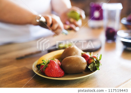 Bowl with fresh fruit for smoothie. Overweight woman in the kitchen cutting fruit on cutting board. 117519443