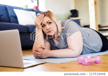 Online fitness training for overweight woman exercising at home, looking at laptop. Workout in living room. Online fitness training for overweight woman exercising at home, looking at laptop. Workout in living room. 117519448