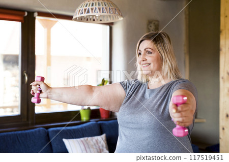Overweight woman exercising at home, using dumbbells. Workout in living room. Overweight woman exercising at home, using dumbbells. Workout in living room. 117519451
