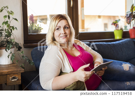 Portrait of beautiful young overweight woman at home, watching tv show on tablet, sitting in armchair. 117519458