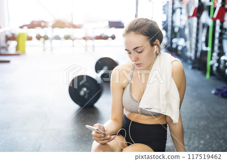 Young fitness girl checking her performance on smatphone after hard workout in gym. Young fitness girl checking her performance on smatphone after hard workout in gym. 117519462
