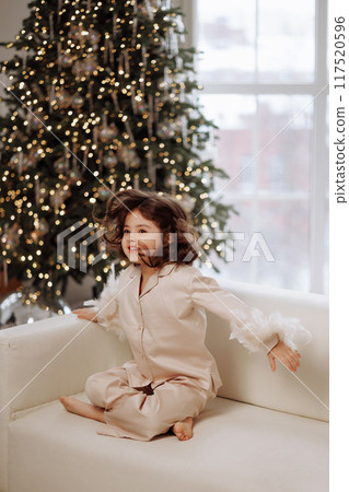 A young girl is sitting on a couch in front of a Christmas tree 117520596