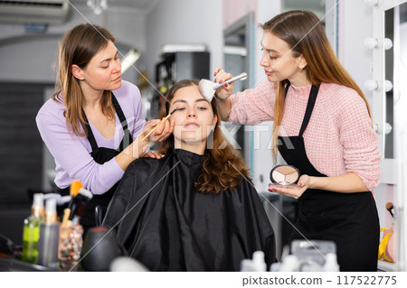 Two makeup artists apply makeup on the face of a young woman Two makeup artists apply makeup on the face of a young woman 117522775