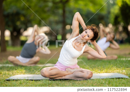 Woman stretching in lotus position at group yoga session in park Woman stretching in lotus position at group yoga session in park 117522894