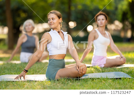 Female yoga instructor stretching in Padmasana pose with group in park 117523247