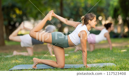 Girl performing Kneeling Bow pose in outdoor group yoga session 117523408