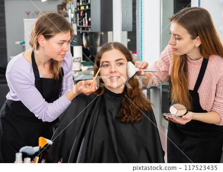 Two makeup artists apply makeup on the face of a young woman 117523435