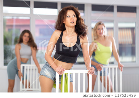 Active female dancer with group of women of different ages practicing strip plastic movements near chair in training hall during dancing classes Active female dancer with group of women of different ages practicing strip plastic movements near chair in training hall during dancing classes 117523459