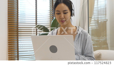 Happy asian woman video call web conference sitting on cozy sofa in living room at home. Smile Female asian people use laptop notebook computer headphones online call working from home watching video 117524823