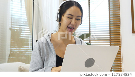 Happy asian woman video call web conference sitting on cozy sofa in living room at home. Smile Female asian people use laptop notebook computer headphones online call working from home watching video 117524824