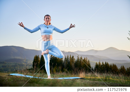 Woman practicing yoga and meditating outdoors in the mountains in a serene, natural setting. Female performing yoga pose on blue mat, with backdrop of beautiful mountain landscape at sunrise or sunset 117525976