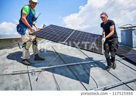 Workers building solar panel system on metal rooftop of house. Two men installers carrying photovoltaic solar module outdoors. Alternative, green and renewable energy generation concept. 117525988