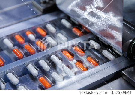 automated medicine packaging machine for orange and white capsules. Pharmaceutical production line with blister packs. Manufacturing and healthcare concept. Close-up shot. 117529641