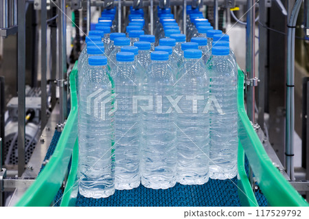 Plastic bottles with blue caps on conveyor belt in water bottling factory. Industrial production line for bottled water manufacturer. Plastic bottles with blue caps on conveyor belt in water bottling factory. Industrial production line for bottled water manufacturer. 117529792
