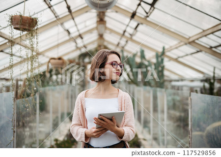 Small greenhouse business. Businesswoman selling flowers and seedlings, standing in greenhouse, looking at camera, smiling. 117529856
