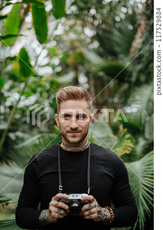 Handsome man with tattoos standing in botanical garden, in the middle of lush green foliage, holding camera and taking photography. 117529884