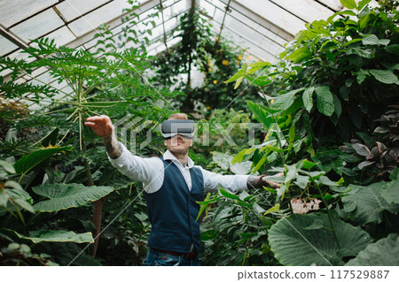 Handsome businessman standing in botanical garden, in the middle of lush green foliage, using VR headset. Handsome businessman standing in botanical garden, in the middle of lush green foliage, using VR headset. 117529887