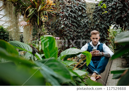 Handsome businessman with smartphone, sitting in botanical garden, in the middle of lush green foliage. Concept of business corporate social responsibility and ESG. Handsome businessman with smartphone, sitting in botanical garden, in the middle of lush green foliage. Concept of business corporate social responsibility and ESG. 117529888