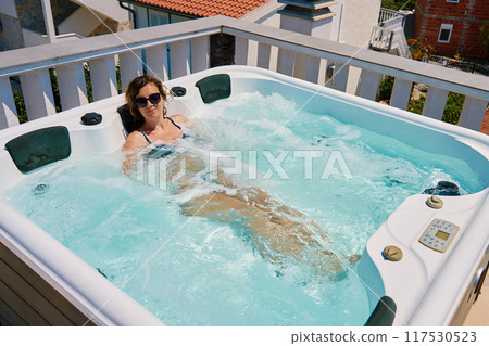 Woman wearing sunglasses and swimsuit relaxing in outdoor hot tub with bubbling water and enjoying sunny day. Female resting in outdoor jacuzzi with beautiful natural landscape Woman wearing sunglasses and swimsuit relaxing in outdoor hot tub with bubbling water and enjoying sunny day. Female resting in outdoor jacuzzi with beautiful natural landscape 117530523