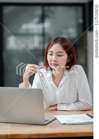 Professional woman in white shirt working on a laptop at a desk with documents, holding glasses, in a modern office setting. 117530656