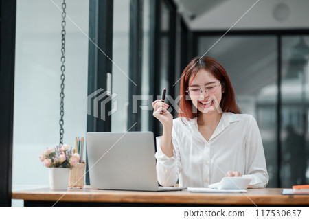 Smiling young woman in white shirt working on a laptop at a modern office desk with natural light and minimalist decor. 117530657