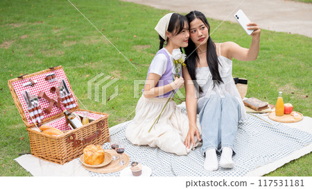 Two lovely young Asian women take a selfie with a smartphone while picnicking together in a park. Two lovely young Asian women take a selfie with a smartphone while picnicking together in a park. 117531181