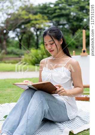A beautiful Asian woman sitting on a picnic blanket in a green park and reading a book. A beautiful Asian woman sitting on a picnic blanket in a green park and reading a book. 117531195