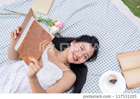 A top view of a positive, lovely Asian woman reading a book and lying on a picnic blanket. A top view of a positive, lovely Asian woman reading a book and lying on a picnic blanket. 117531205