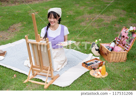 A pretty Asian woman is painting on a canvas easel while sitting on a picnic blanket in a park. A pretty Asian woman is painting on a canvas easel while sitting on a picnic blanket in a park. 117531206