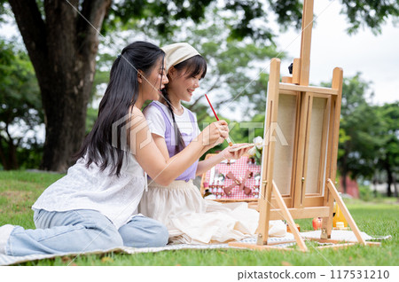 Two positive and lovely young Asian women are enjoying painting on a canvas easel together in a park 117531210