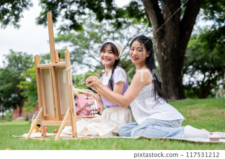 Two pretty Asian women are sitting together on a picnic blanket with a canvas easel in a park. Two pretty Asian women are sitting together on a picnic blanket with a canvas easel in a park. 117531212