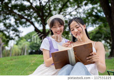 Two beautiful and positive Asian friends are reading a book together while picnicking in a park. Two beautiful and positive Asian friends are reading a book together while picnicking in a park. 117531225