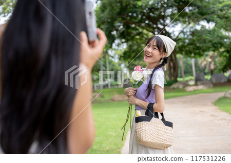 Lovely, smiling Asian girl is having her photo taken by a friend while strolling together in a park. Lovely, smiling Asian girl is having her photo taken by a friend while strolling together in a park. 117531226