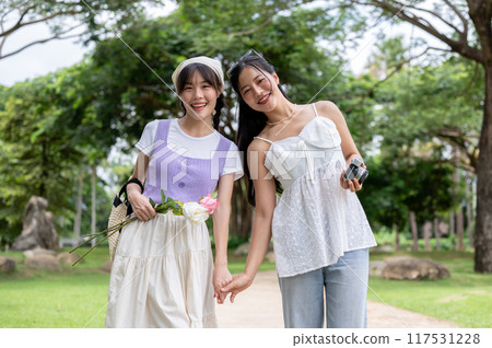 Two lovely Asian women are holding hands, strolling through a park, and smiling at the camera. 117531228