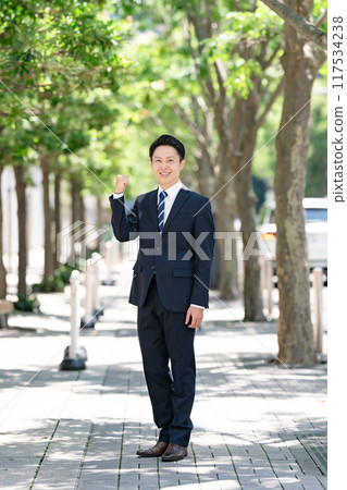 Businessman standing on a tree-lined street, fist pump Businessman standing on a tree-lined street, fist pump 117534238