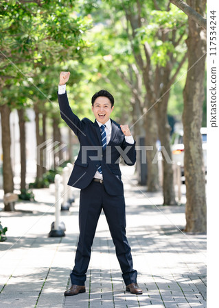 Businessman standing on a tree-lined street, fist pump Businessman standing on a tree-lined street, fist pump 117534244