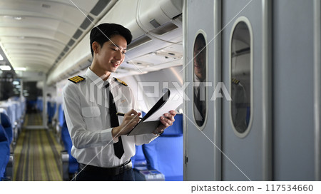 Smiling asian pilot standing inside an airplane, holding clipboard and reviewing or completing some paperwork Smiling asian pilot standing inside an airplane, holding clipboard and reviewing or completing some paperwork 117534660