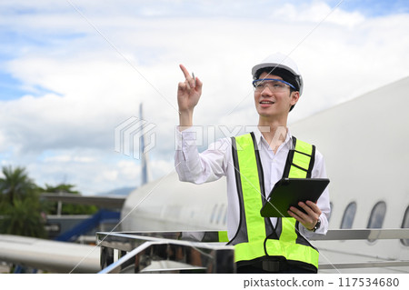 Young aviation engineer with digital tablet standing on a metal railing next to an airplane 117534680
