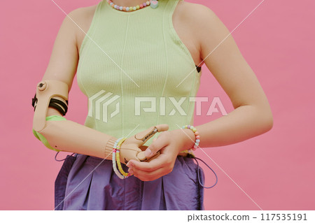 Midsection of unrecognizable teen girl with arm prosthesis wearing stylish clothes posing for camera in studio, pink background 117535191