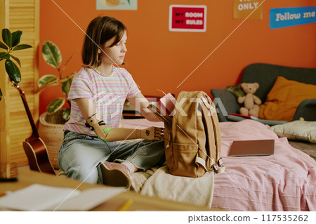 Girl with disability in casual clothes sitting on bed packing her backpack before going to school, medium long shot 117535262