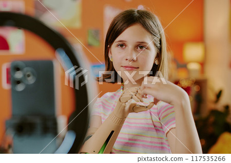 Gen alpha teen influencer with arm prosthesis showing hand heart while smartphone camera on tripod recording video Gen alpha teen influencer with arm prosthesis showing hand heart while smartphone camera on tripod recording video 117535266