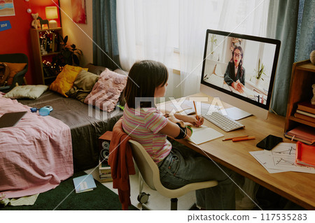 Modern teen girl with prosthetic arm sitting at desk and looking at screen while having online lesson with school teacher 117535283